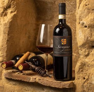 Bottiglia di vino Sagrantino Saragano con calice e tappi su una mensola di pietra.